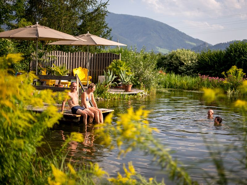 Zwei erwachsene Campinggäste, die auf einem Steg am Wasser sitzen und ihre Beine ins Wasser hängen haben. Im Hintergrund Berge